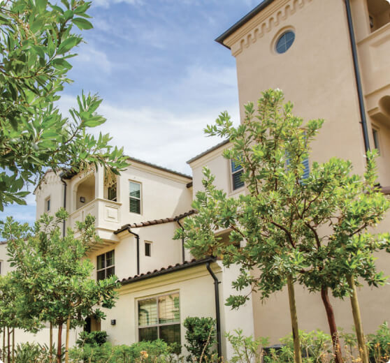 Mediterranean-style residential building with beige stucco walls, terracotta roof tiles, and lush trees in the foreground.