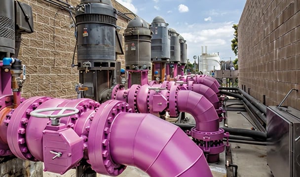 Outdoor industrial piping system with large purple pipes, valves, and gray storage tanks along brick walls.