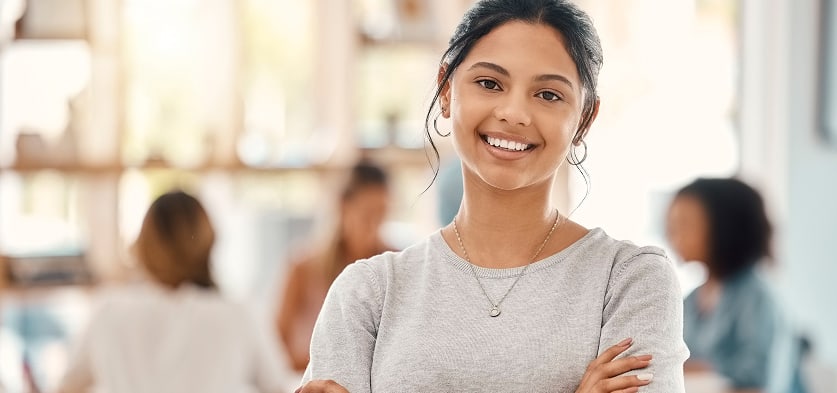 Smiling woman with folded arms in a bright, modern workspace with blurred colleagues in the background.