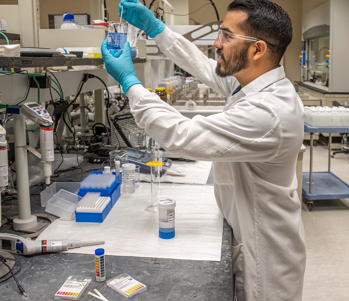 Scientist in a white lab coat and safety goggles pipettes a blue liquid from a beaker in a busy laboratory.