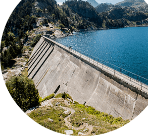 Concrete dam angled along a blue lake with pine trees and rocky hills in the background