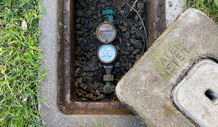 Water meter in an open rectangular utility pit surrounded by grass, with gravel and a valve visible inside.
