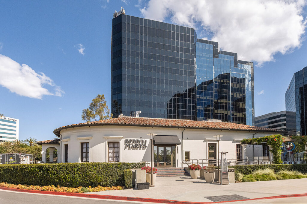 White circular building with a terracotta roof in front of modern glass skyscrapers under a blue sky.