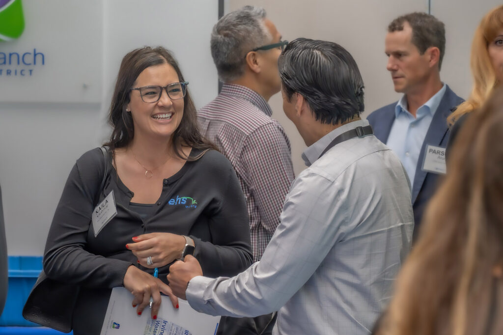 Smiling woman wearing glasses and a black cardigan greets a man in a light shirt at a networking event, with others chatting in the background.