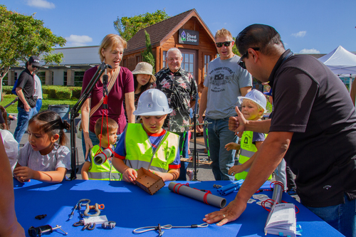 Outdoor workshop with kids wearing safety vests gathered around a blue tool table as a man demonstrates.