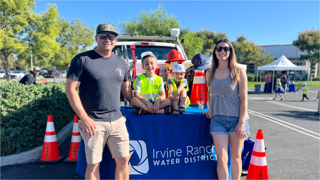 Family posing at Irvine Ranch Water District safety booth with kids sitting on a table and safety gear nearby.