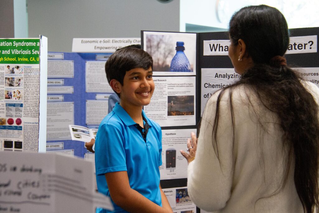 Smiling boy in a blue polo explains a science fair poster to an adult mentor.