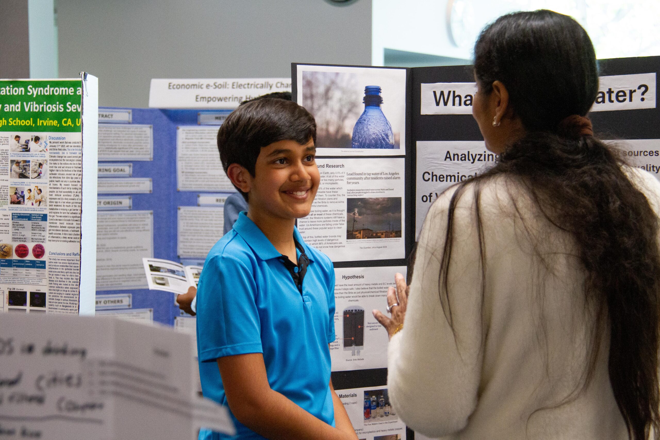 Smiling boy in a blue polo explains a science fair poster to an adult mentor.