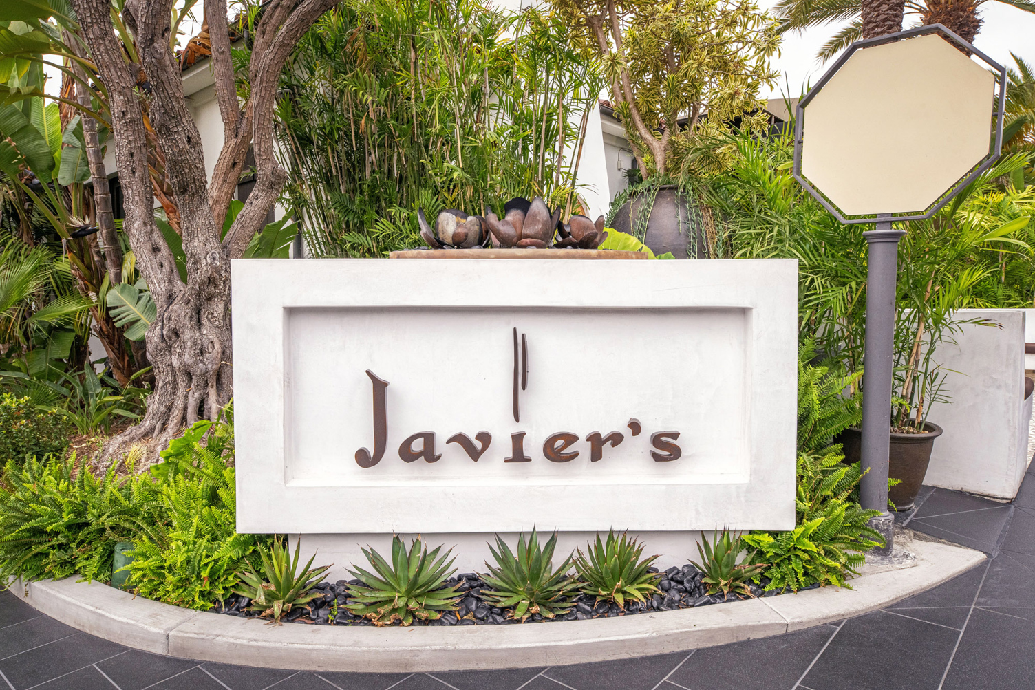 White rectangular sign reading Javier's, surrounded by tropical plants and stone landscaping.