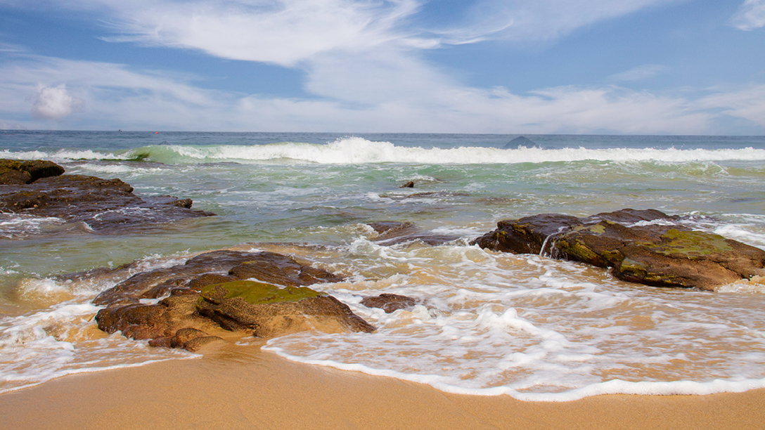 Sandy beach with large rocks in the foreground, turquoise waves and a clear blue sky with wispy clouds.