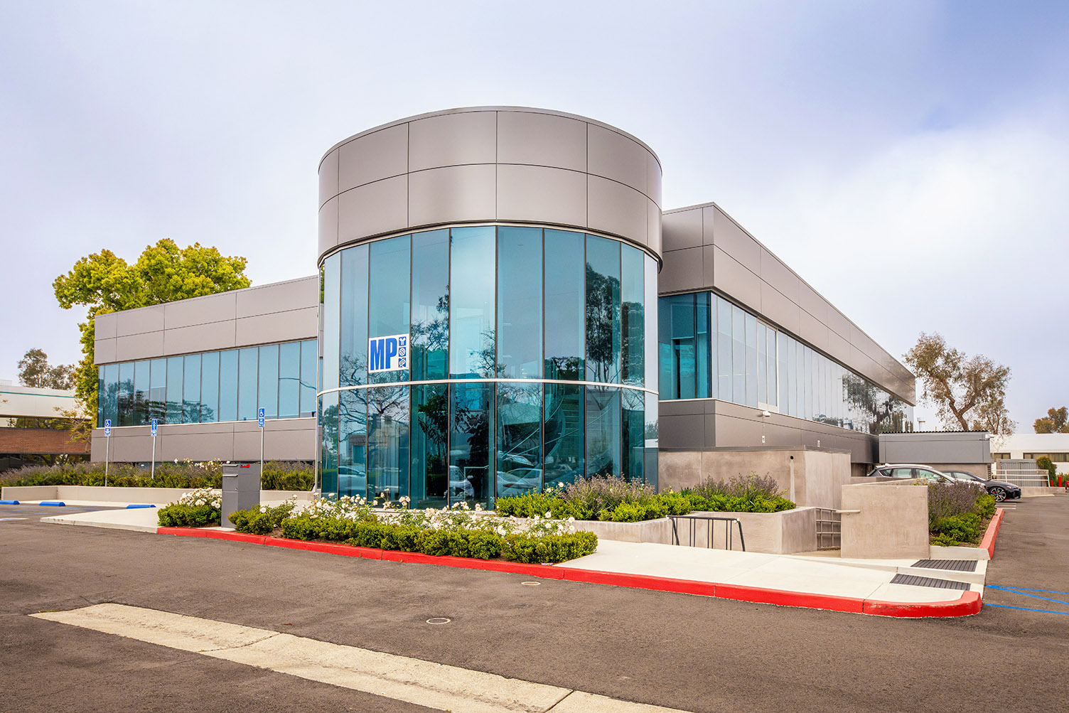 Modern corporate office building with glass facade and curved cylindrical tower, surrounded by greenery and a parking lot.