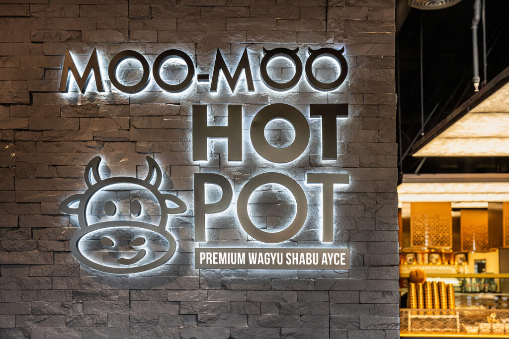 Moo-Moo Hot Pot sign with backlit letters and a cow logo on a textured stone wall