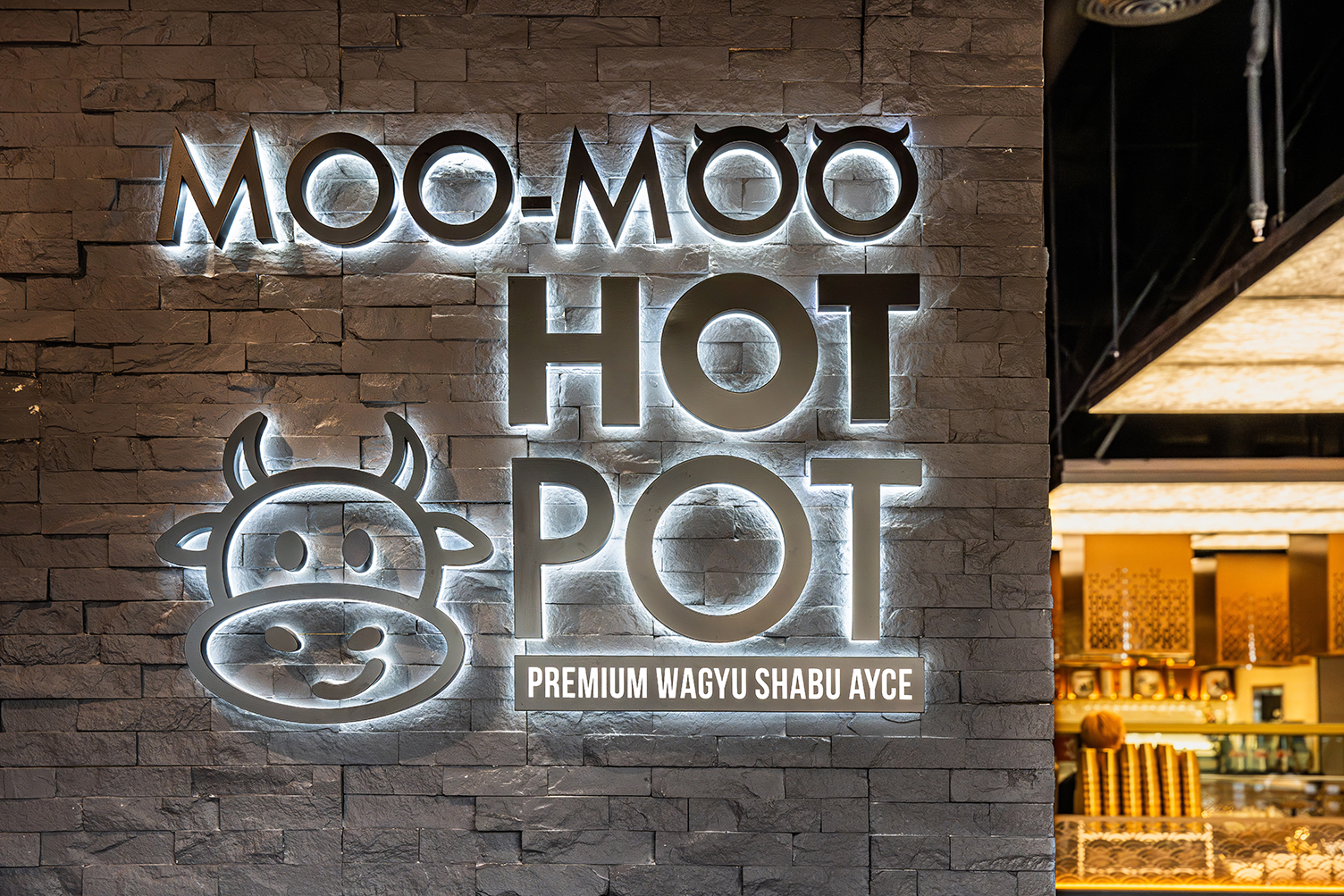 Moo-Moo Hot Pot sign with backlit letters and a cow logo on a textured stone wall