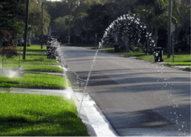 Residential street with water jets spraying arcing streams across the road beside green lawns.