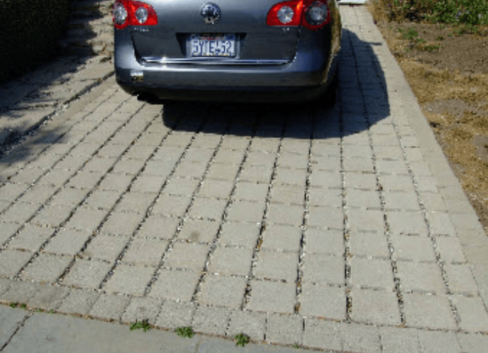 Rear view of a blue sedan parked on a driveway paved with rectangular concrete blocks in a grid pattern.