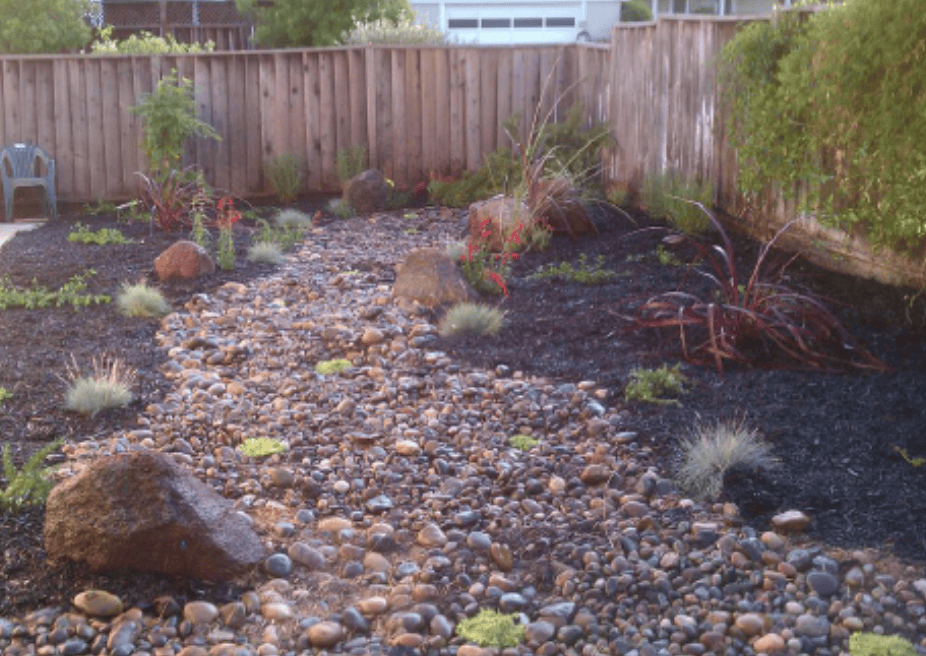 Backyard landscape with a rocky dry creek bed winding through mulch and drought-tolerant plants, wooden fence in back