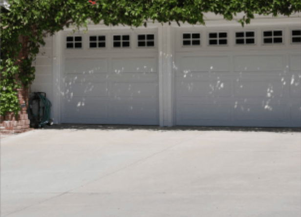 Two-car garage with white sectional doors, small square windows along the top, and a concrete driveway under a leafy canopy.