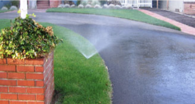 Sprinkler watering a curved lawn beside a brick planter in a suburban front yard