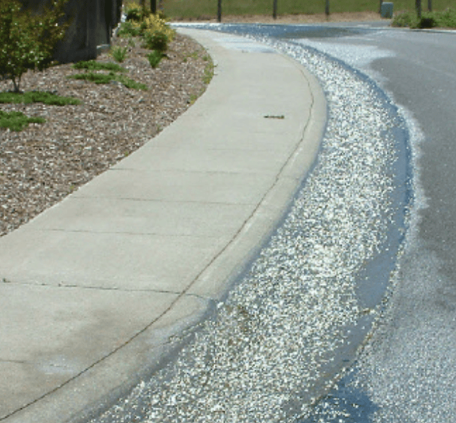 Curved concrete sidewalk beside a landscaped strip and a street with a narrow band of white gravel along the curb.