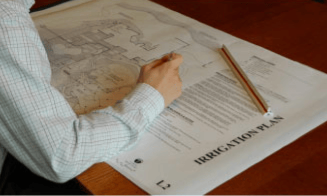 Person marking details on an irrigation plan blueprint spread on a wooden desk