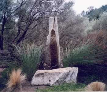 Tall slender stone sculpture with an arched central opening on a raised pedestal, set among grasses in a park.