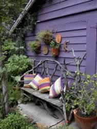 Purple wooden shed exterior with a black metal bench dressed in colorful striped cushions, surrounded by greenery.