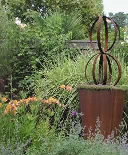 Rusty metal sculpture with looping circular rings in a lush garden with orange flowers and tall grasses.