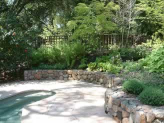 Backyard garden with a curved stone retaining wall, lush shrubs, and a wooden fence.