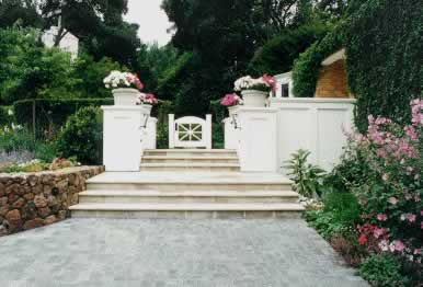 White garden entrance with steps to a decorative gate, flanked by planters and blooming flowers.