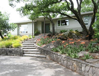 Residential front yard with terraced stone garden beds, concrete steps, and a mid-century house entrance.