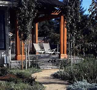 Wooden pergola over a deck with white lounge chairs beside a garden path.