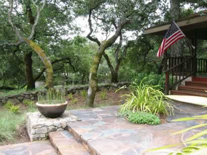 Stone patio with steps, curved vine-covered arch, potted plant, and an American flag by a wooden porch in a garden.