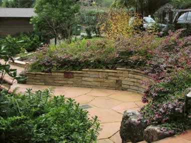 Curved stone retaining wall with flowering shrubs and a paved patio in a residential garden.