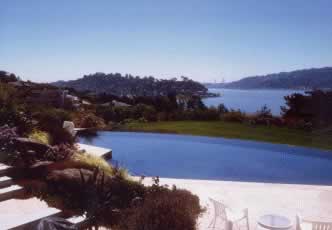Outdoor pool with white decking perched on a hillside, overlooking a blue bay and distant coastline.