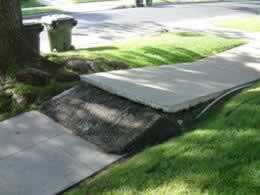 Residential sidewalk with a curb ramp partially covered by a white tarp and two green trash cans nearby.