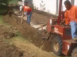 Two construction workers in orange shirts dig and level soil beside a small red skid-steer loader against a white wall.