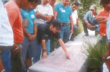 Group of people gathered around a rectangular slab on the ground, one person leaning over to touch or inspect it outdoors.