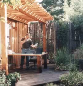 Person wearing an apron grills at an outdoor wooden deck under a pergola in a garden.
