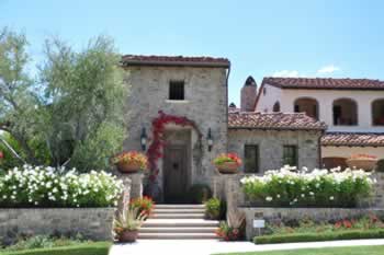 Stone villa with red-tile roof, arched doorway, and flower-filled path under a bright blue sky.