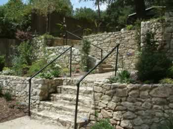 Stone staircase with black metal handrails amid terraced stone walls and green plants.