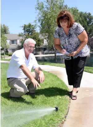 Old man kneeling on a suburban lawn beside a sprinkler; a woman stands nearby holding a clipboard.